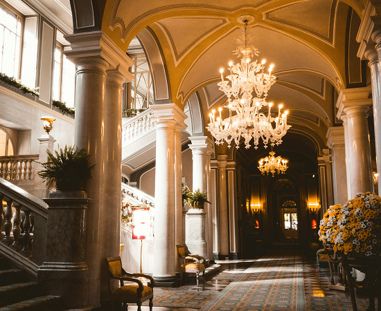 Elegante Hotellobby mit hohen Decken, Säulen und Kronleuchtern. Eine geschwungene Treppe führt nach oben, während bunte Blumenarrangements und Sitzgelegenheiten den Raum schmücken. Tageslicht strömt durch große Fenster.