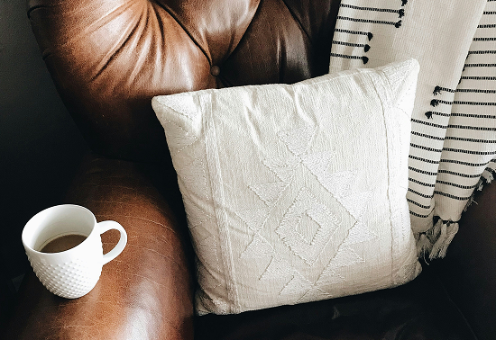 Gemütliche Ecke eines Sessels mit einem hellen Kissen, einer Tasse Kaffee und einer gestreiften Decke im Hintergrund.